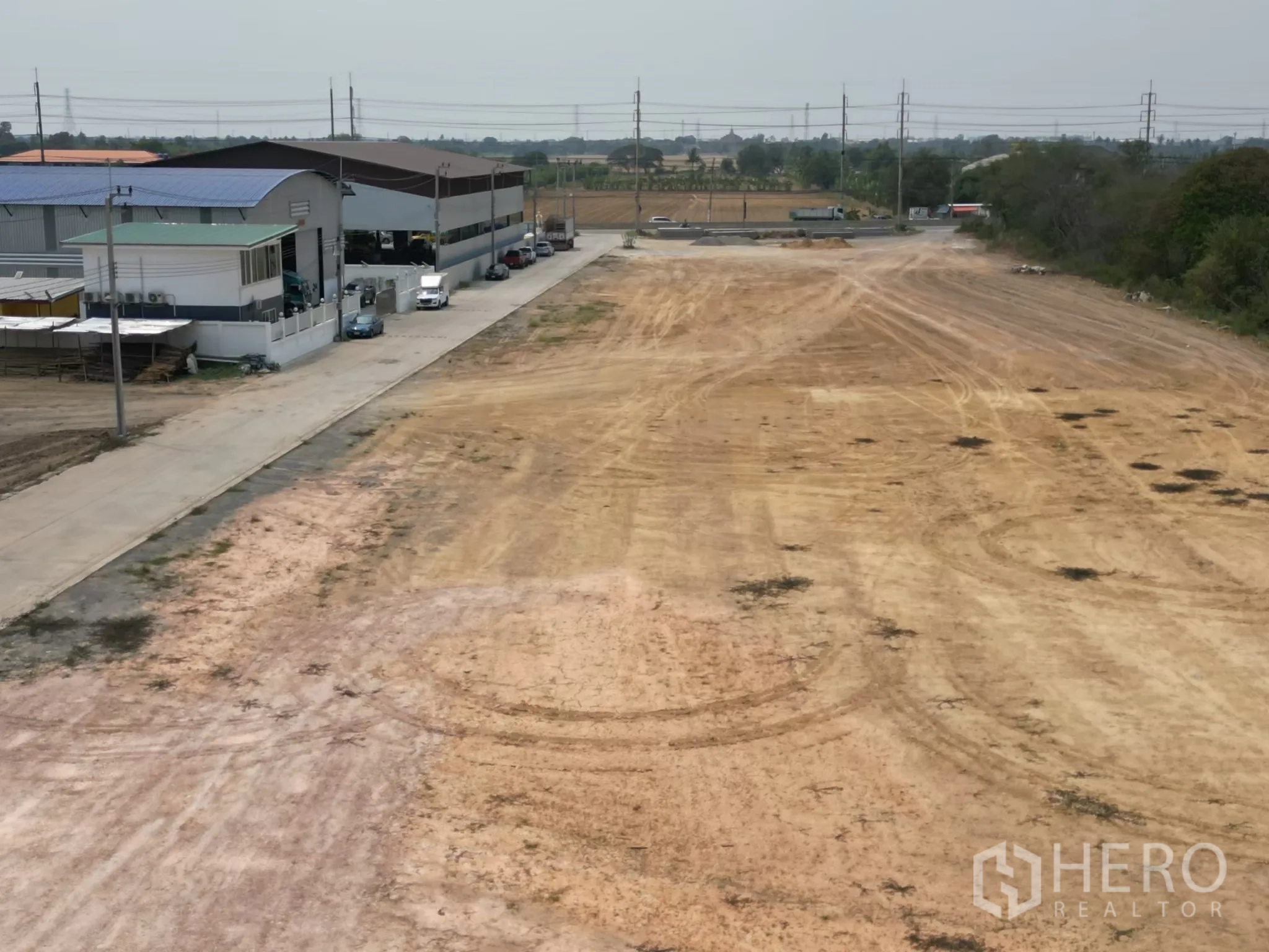 Land for sale in Sai Noi, Nonthaburi - Cleared industrial land beside a paved access road with nearby warehouses and utility poles.