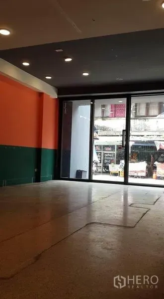 店屋 出租在Bang Khen, Bangkok - 一层零售空间，宽幅玻璃门面，橙绿双拼墙面与嵌灯，临街视野。