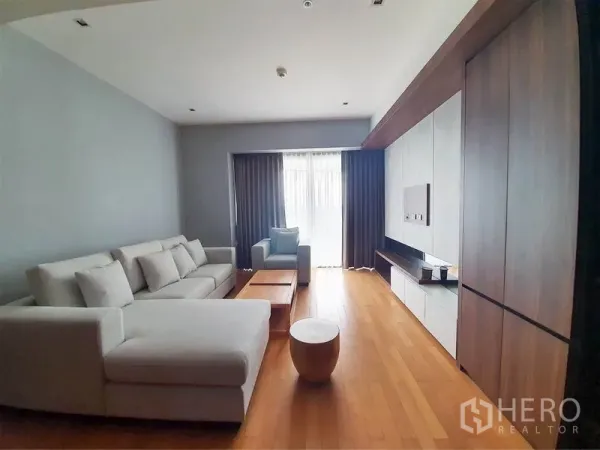 Condominium for rent in Yan Nawa, Bangkok - Bright living room with L-shaped sofa, wood floors and TV wall facing a curtained balcony.