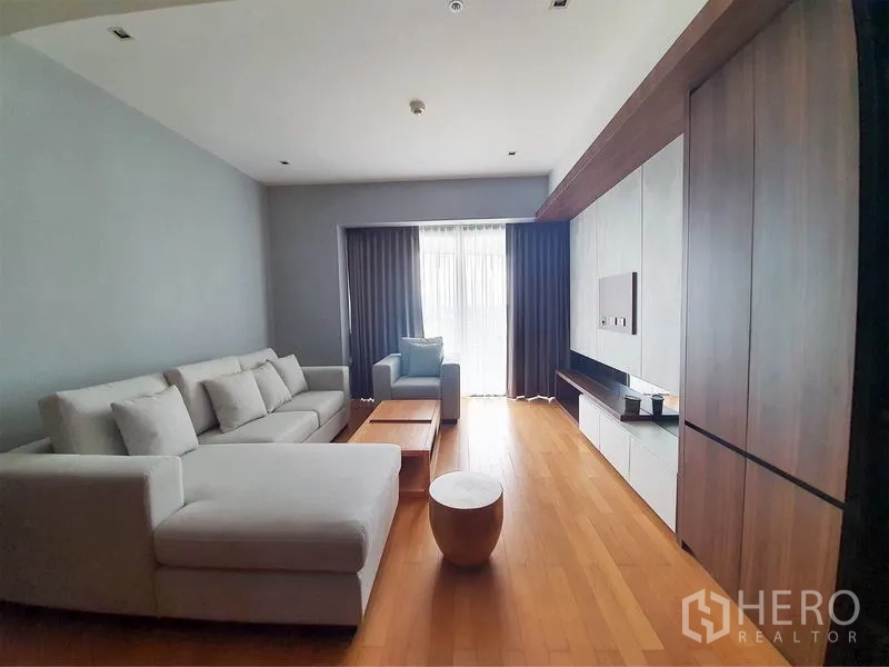 Condominium for rent in Yan Nawa, Bangkok - Bright living room with L-shaped sofa, wood floors and TV wall facing a curtained balcony.