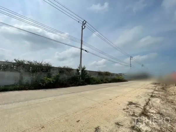 Land for sale in Sai Noi, Nonthaburi - Wide concrete access road with power lines and a perimeter wall beside the plot.