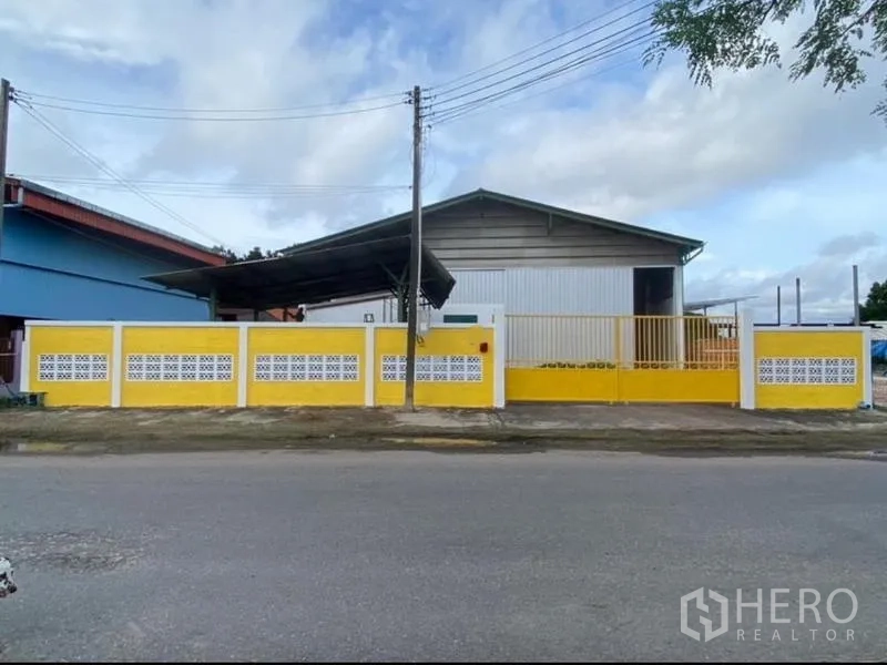 Warehouse for rent in Mueang Rayong, Rayong - Wide street-facing elevation of the warehouse behind a yellow wall and gate.