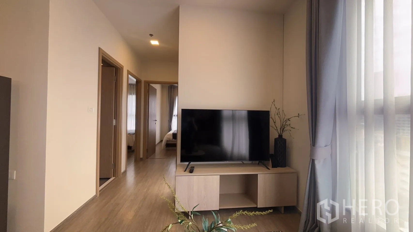 Condominium for rent in Watthana, Bangkok - Living area with TV on a low console and hallway opening to the bedroom, lit by a large curtained window.