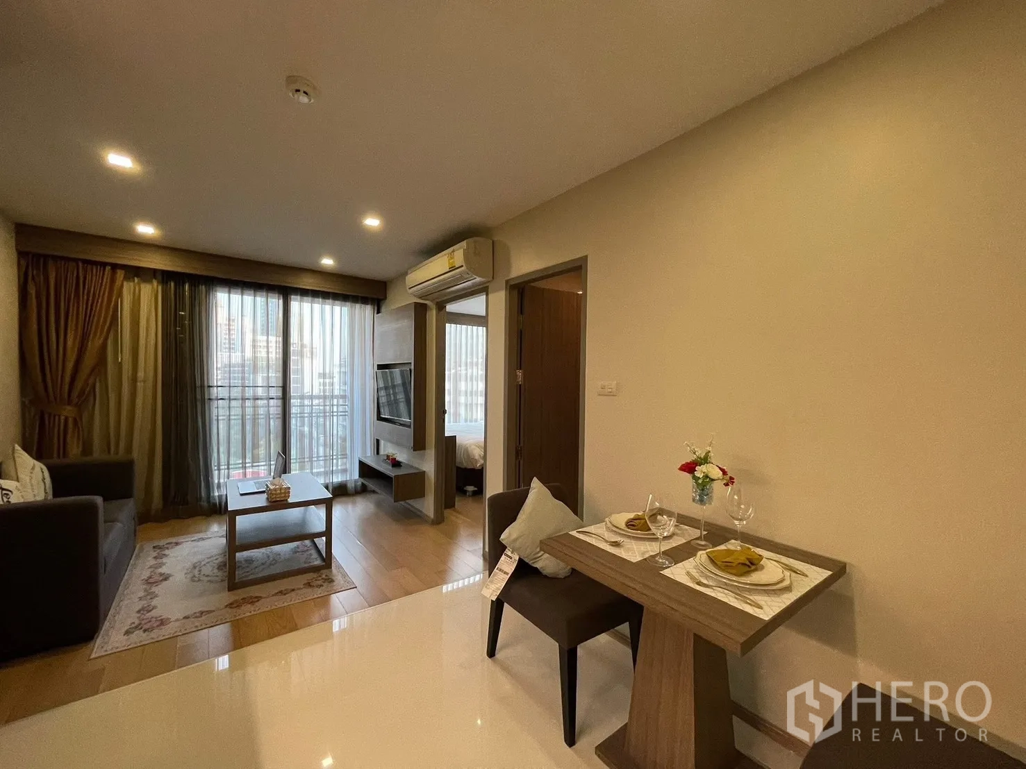 Condominium for rent in Watthana, Bangkok - Living room with balcony doors, wall-mounted TV, and a compact dining table set for two.