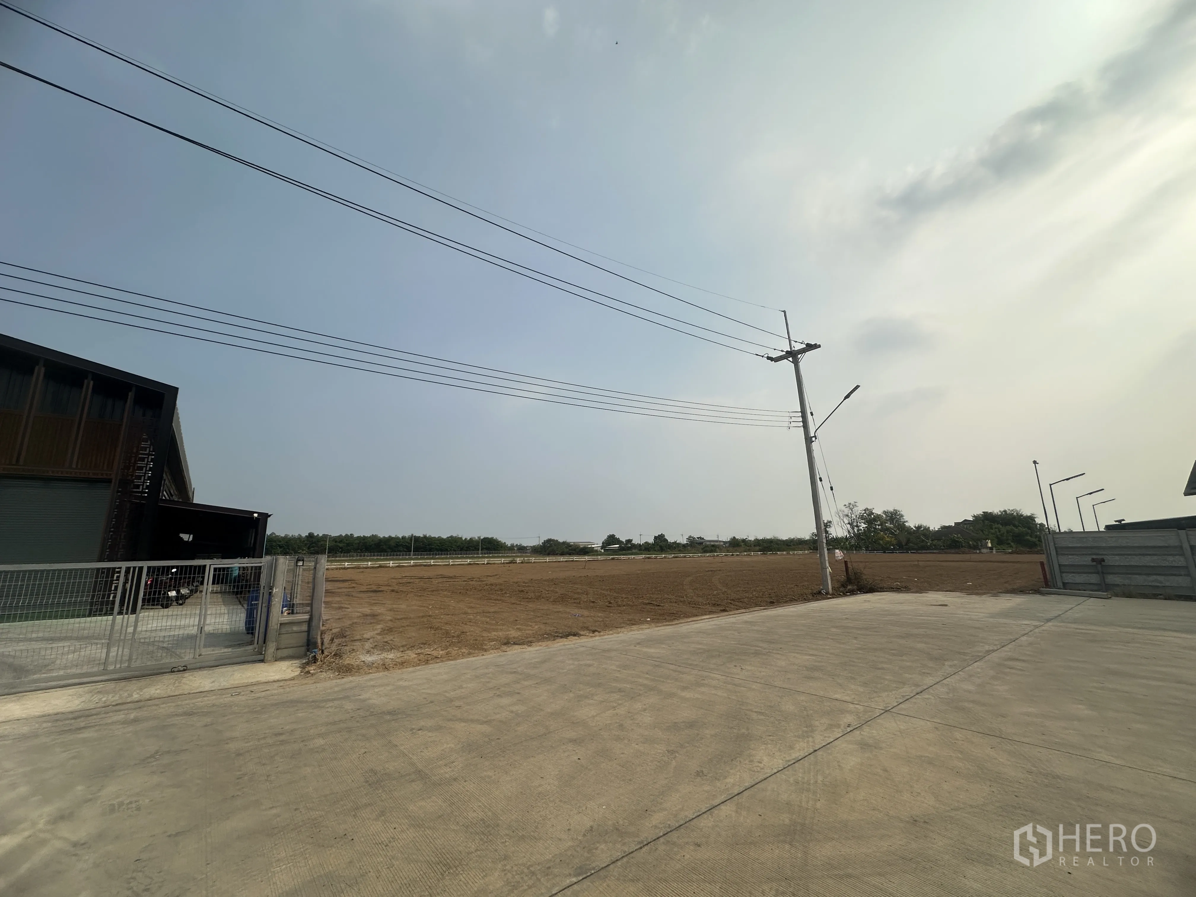 土地 for sale in Lam Luk Ka, Pathum Thani - 从另一角度拍摄的空地，旁有带围栏的建筑，沿混凝土通道设路灯与电缆。