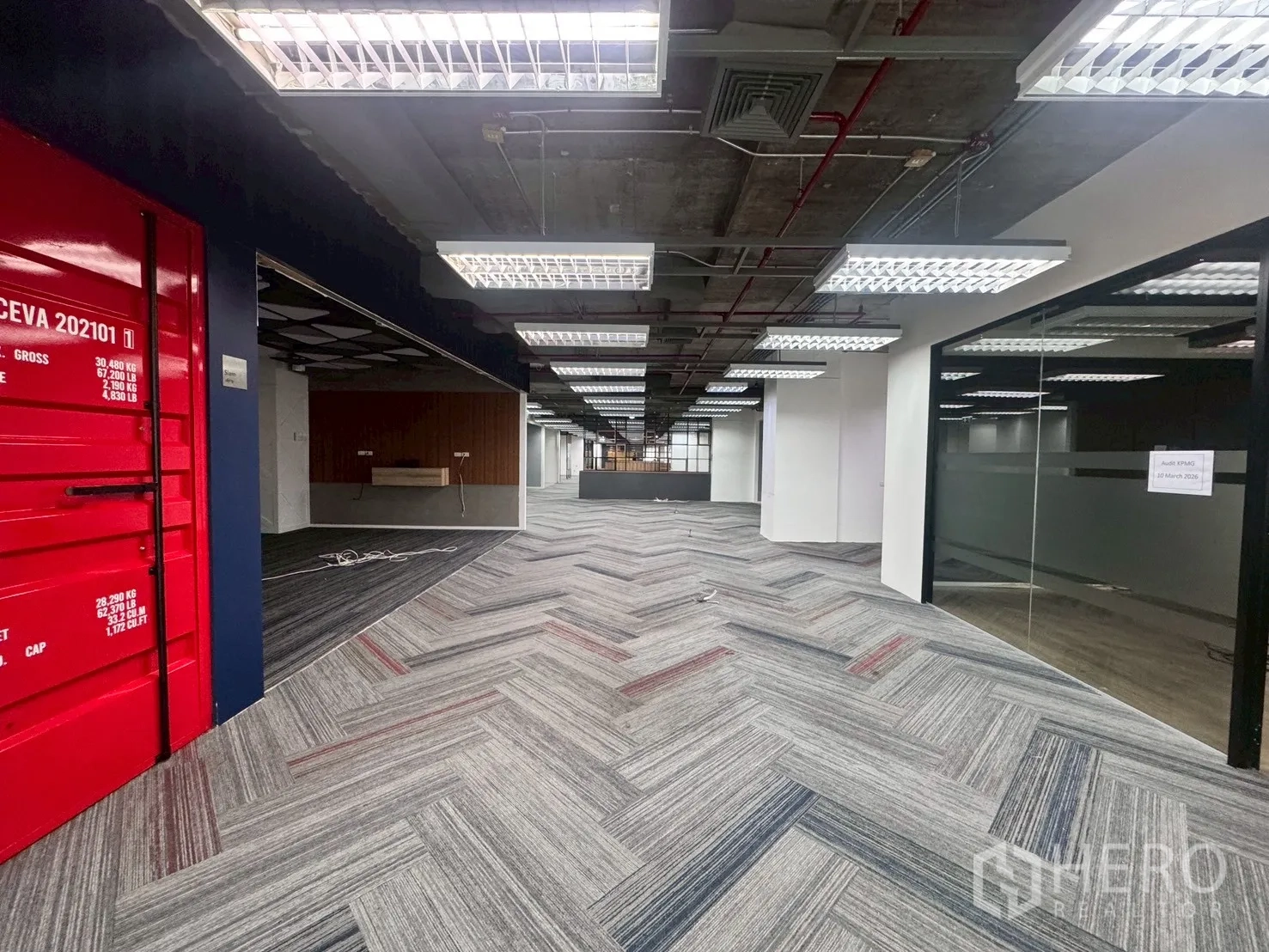 办公室 for rent in Watthana, Bangkok - 中轴通道带红色集装箱风格装饰，右侧为玻璃房与网格灯顶。