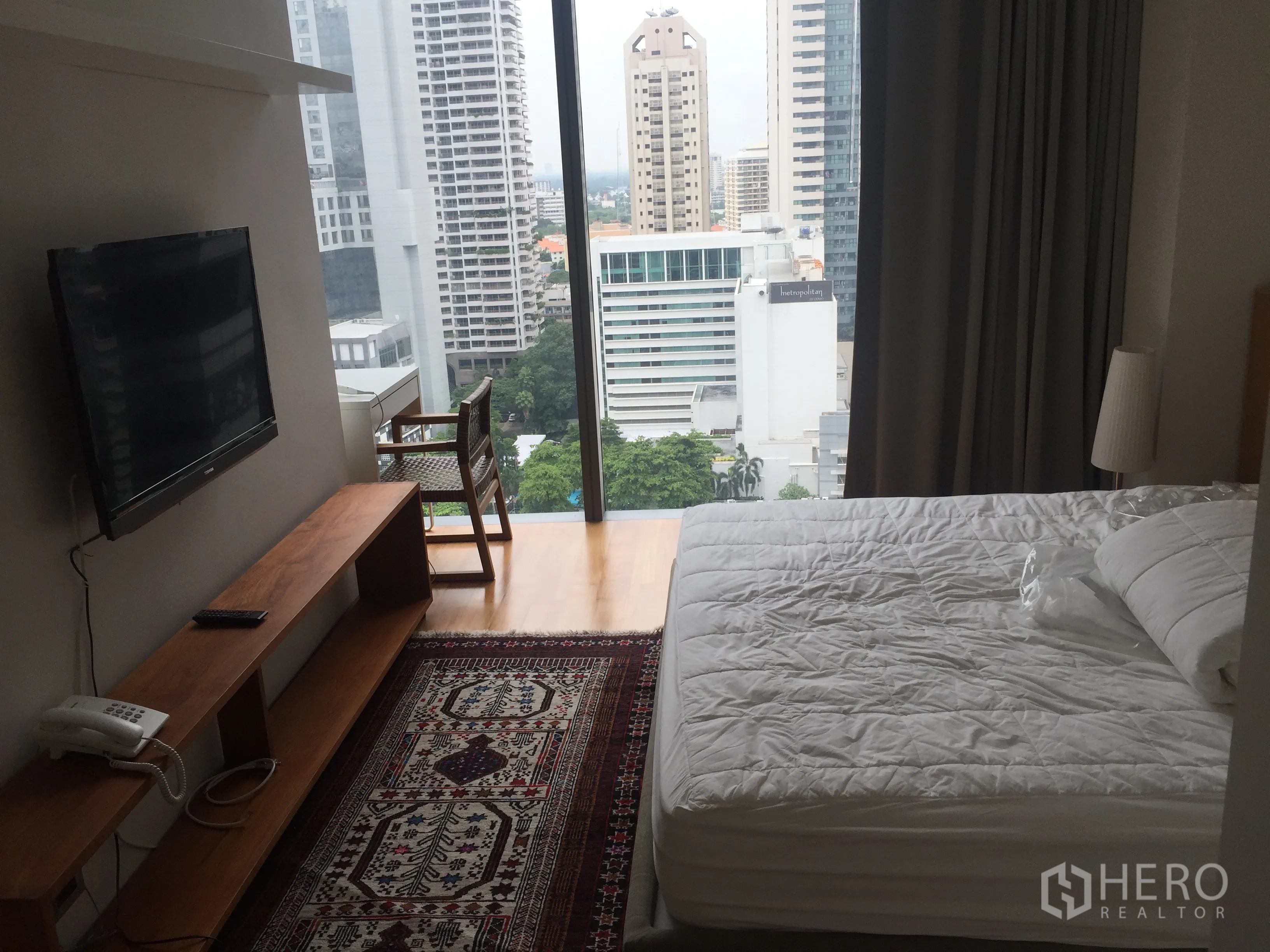 公寓 出租在Bang Rak, Bangkok - 主卧落地窗观景，白色床铺与图案地毯，享城市视野。