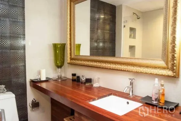 Condominium for rent in Watthana, Bangkok - Bathroom with long wooden vanity, under-mount sink, ornate gold mirror and textured tile accent wall.