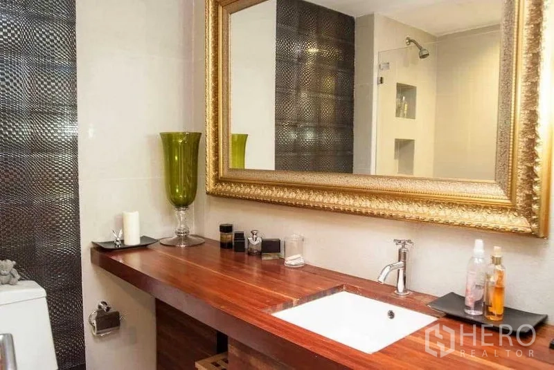 Condominium for rent in Watthana, Bangkok - Bathroom with long wooden vanity, under-mount sink, ornate gold mirror and textured tile accent wall.