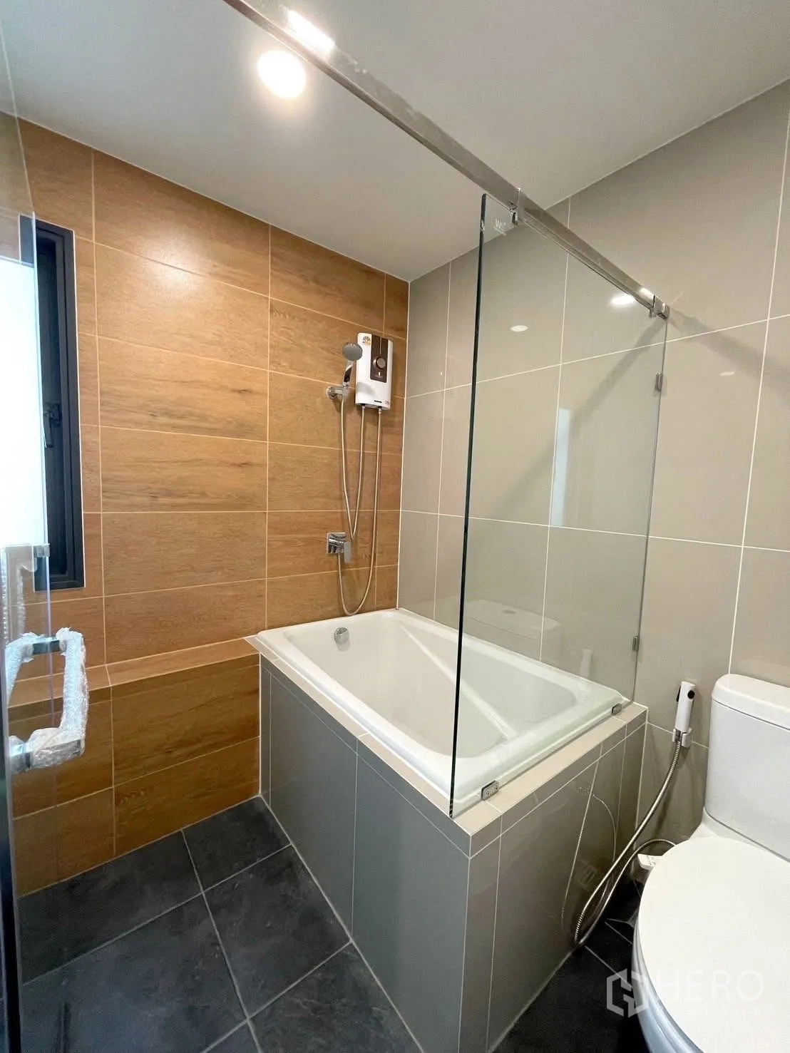 Detached House for rent in Bang Phli, Samut Prakan - Modern bathroom with bathtub, glass shower screen, wood-look wall tiles, and a water heater.