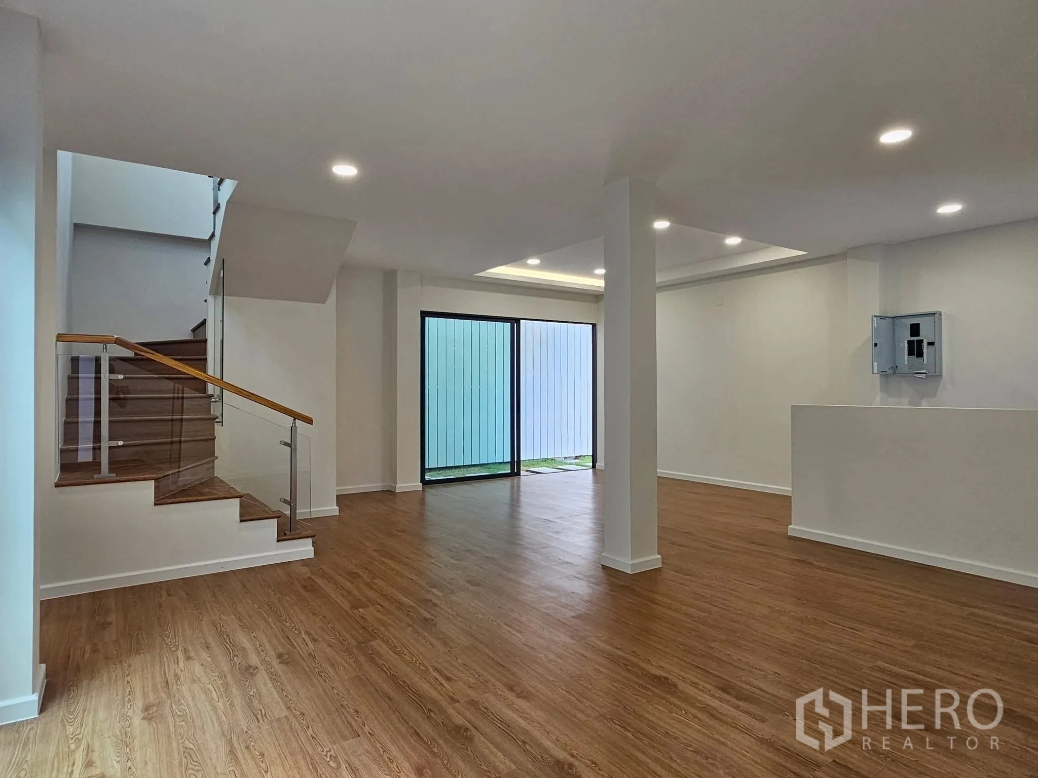 บ้านเดี่ยว for sale in Lat Phrao, Bangkok - โถงรับแขกแบบโอเพ่น พื้นลายไม้ ไฟดาวน์ไลท์ พร้อมบันไดราวกระจกและประตูกระจกสไลด์เปิดสู่คอร์ทยาร์ด