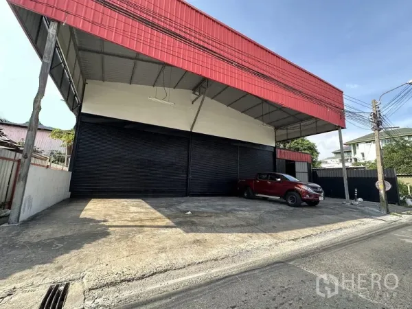 คลังสินค้า ให้เช่าในLat Phrao, Bangkok - ด้านหน้าโกดังลาดพร้าว โครงหลังคาใหญ่สีแดง พร้อมลานขนถ่ายสินค้าและประตูม้วนสีดำหลายบาน