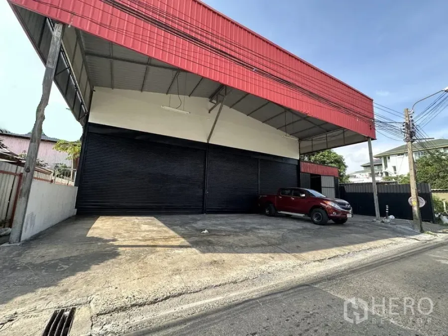 คลังสินค้า ให้เช่าในLat Phrao, Bangkok - ด้านหน้าโกดังลาดพร้าว โครงหลังคาใหญ่สีแดง พร้อมลานขนถ่ายสินค้าและประตูม้วนสีดำหลายบาน