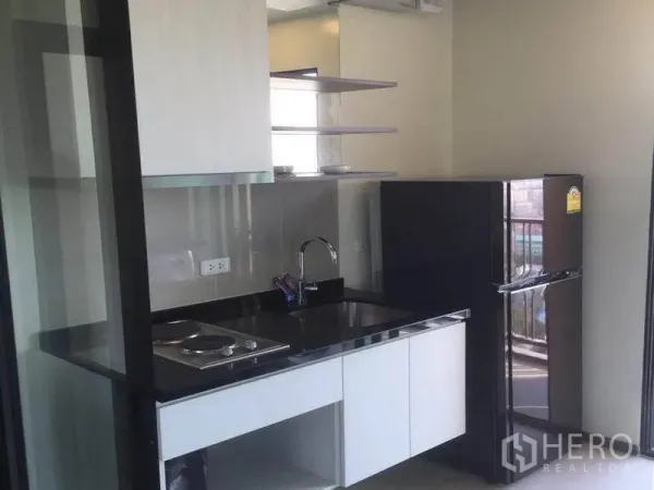 Condominium for sale in Watthana, Bangkok - Compact kitchenette with black countertop, electric hob, sink, open shelves and a refrigerator behind sliding glass.