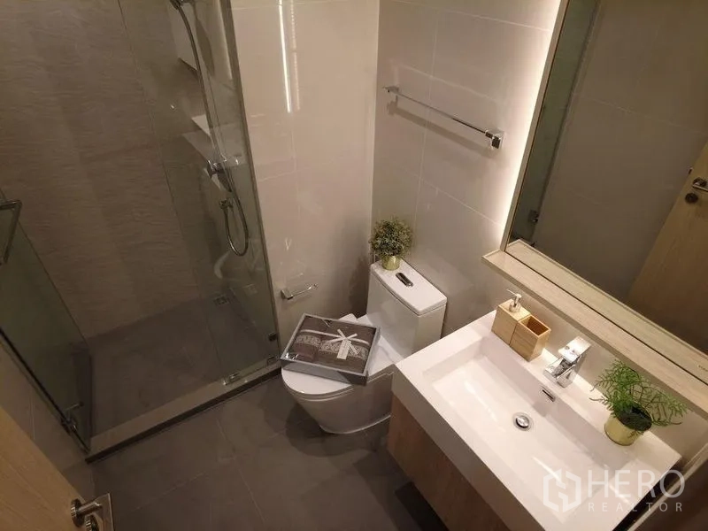 Condominium for sale in Watthana, Bangkok - Modern bathroom with glass-enclosed shower, vanity, and toilet in neutral tones.