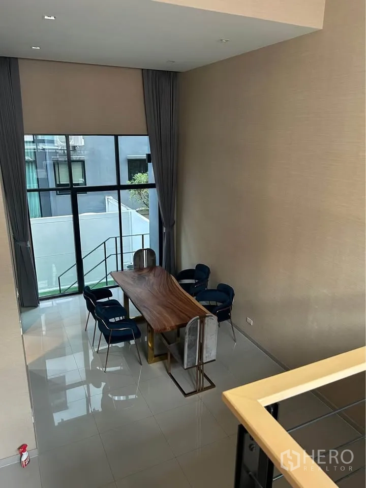 Townhouse for rent in Suan Luang, Bangkok - Double-height dining area with floor-to-ceiling windows and a live-edge wooden table with blue chairs.