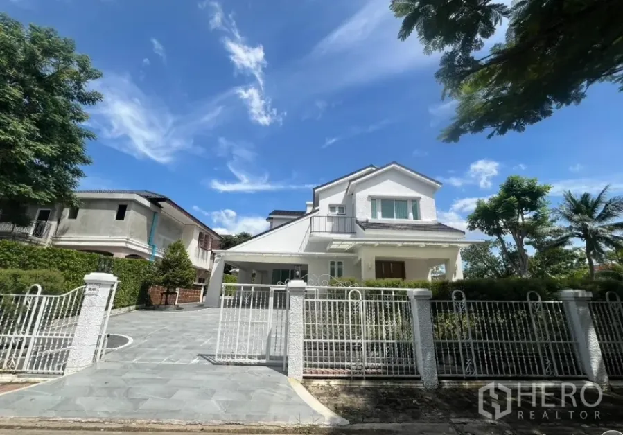 独立式住宅 出租在Mueang Samut Prakan, Samut Prakan - 白色两层独栋住宅正面，带大门和车道，蓝天之下。