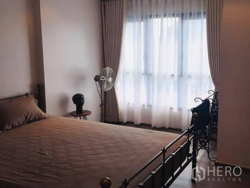 Condominium for sale in Watthana, Bangkok - Cozy bedroom with metal bed frame, sheer curtains, standing fan and soft natural light.