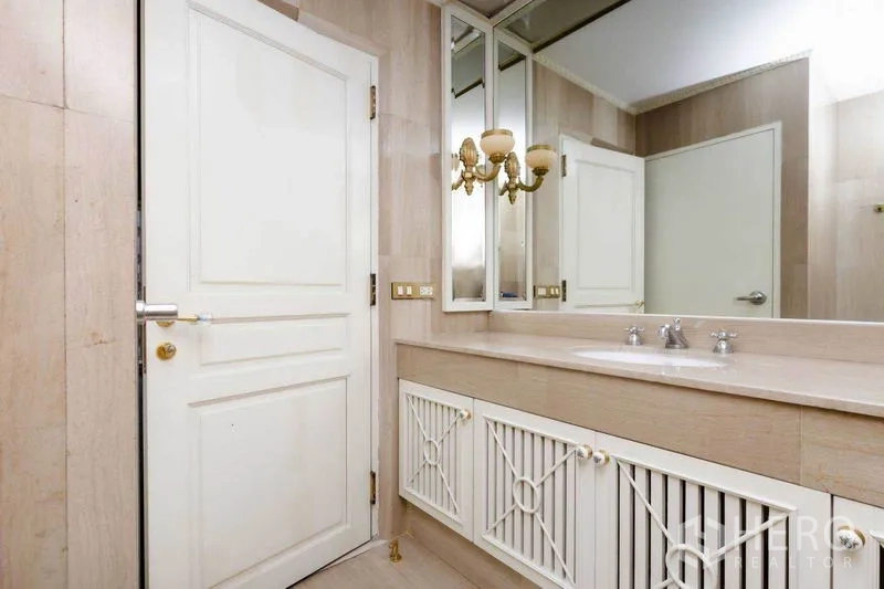 Condominium for rent or sale in Pathum Wan, Bangkok - Classic beige-tiled bathroom with long vanity, mirrored cabinets, and brass wall sconce.