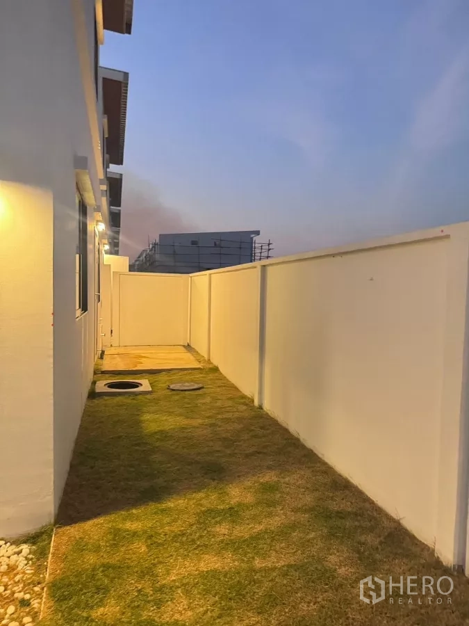 Detached House for rent in Samut Prakan - Long side garden with grass path and a tall white privacy wall behind the house.