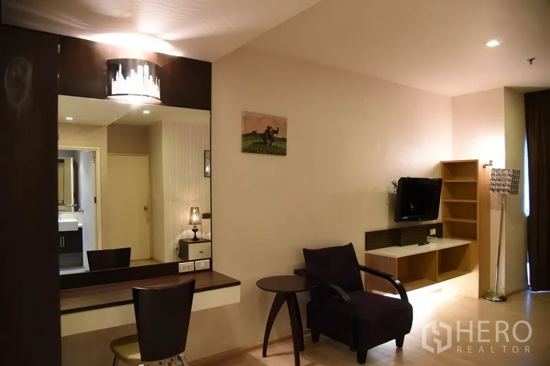 Condominium for rent in Khlong Toei, Bangkok - Living area with wall-mounted TV unit, vanity desk with large mirror, lounge chair and access to bathroom.