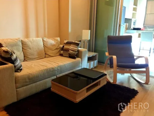 Condominium for rent in Ratchathewi, Bangkok - Cozy living room with beige sofa, striped cushions, rocking chair and glass partition on warm wood floors.