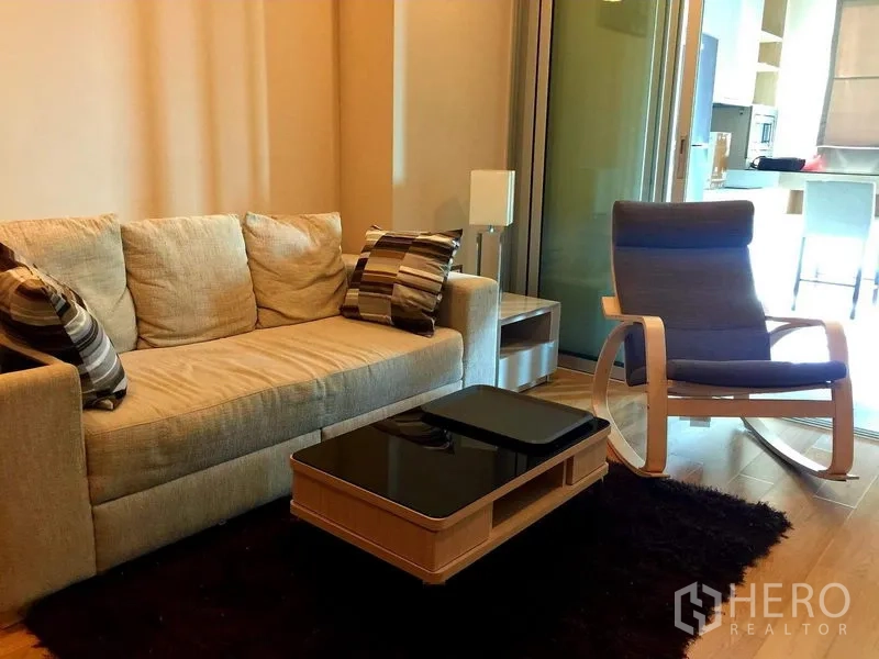 Condominium for rent in Ratchathewi, Bangkok - Cozy living room with beige sofa, striped cushions, rocking chair and glass partition on warm wood floors.