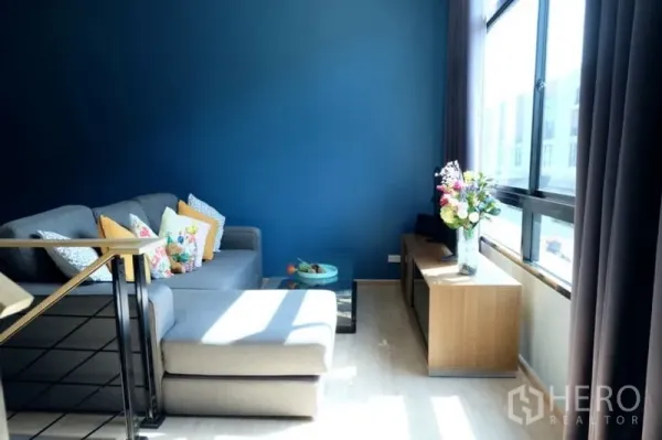 Townhouse for rent in Khlong Toei, Bangkok - Sunlit living room with grey sectional sofa, blue feature wall and large windows.