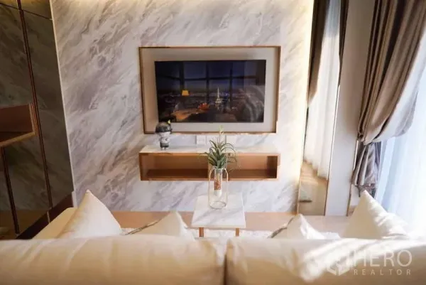 Condominium for rent or sale in Watthana, Bangkok - Cozy living room with a marble feature wall, wall-mounted TV, floating shelf, and floor-to-ceiling curtains.