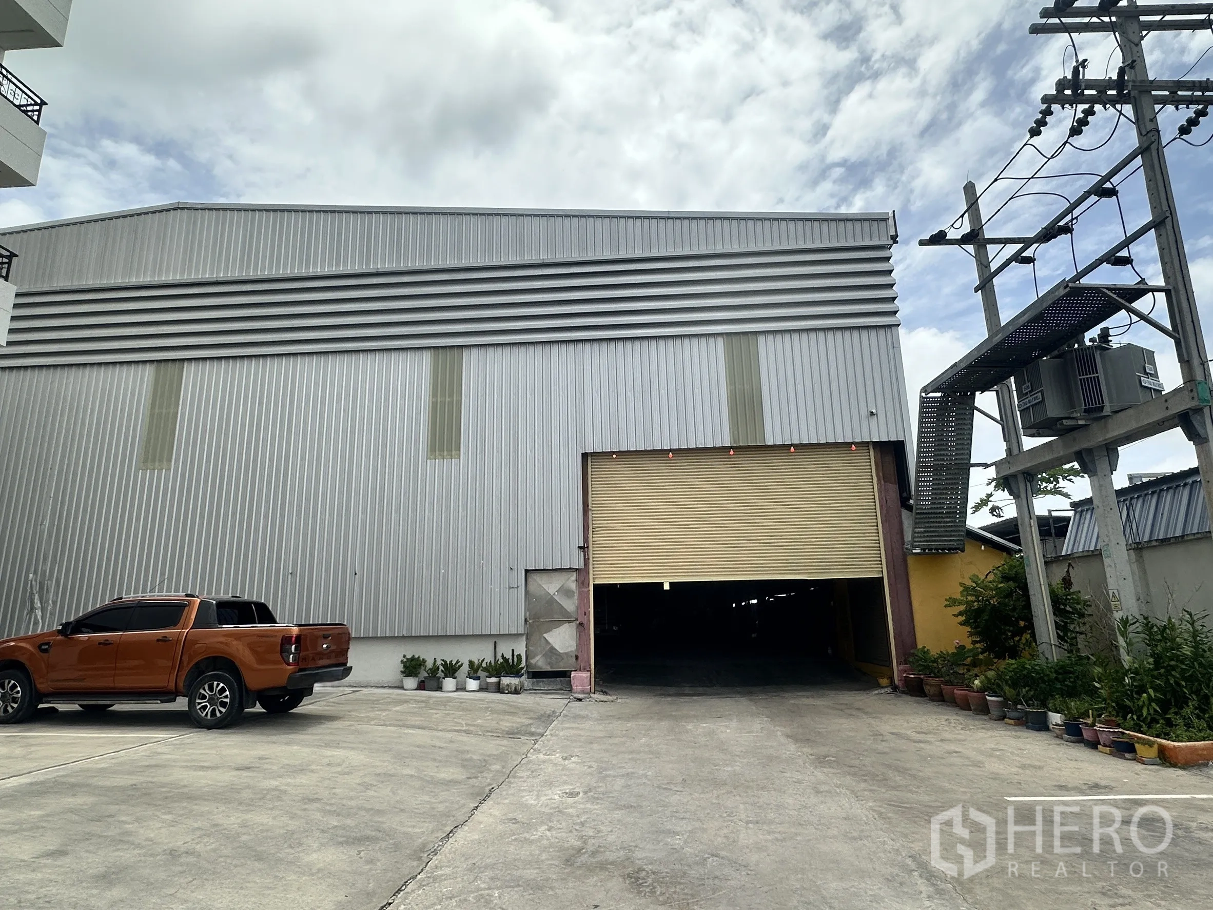 Warehouse for rent in Mueang Samut Prakan, Samut Prakan - Metal-clad warehouse exterior with wide roller shutter, driveway and power transformer.