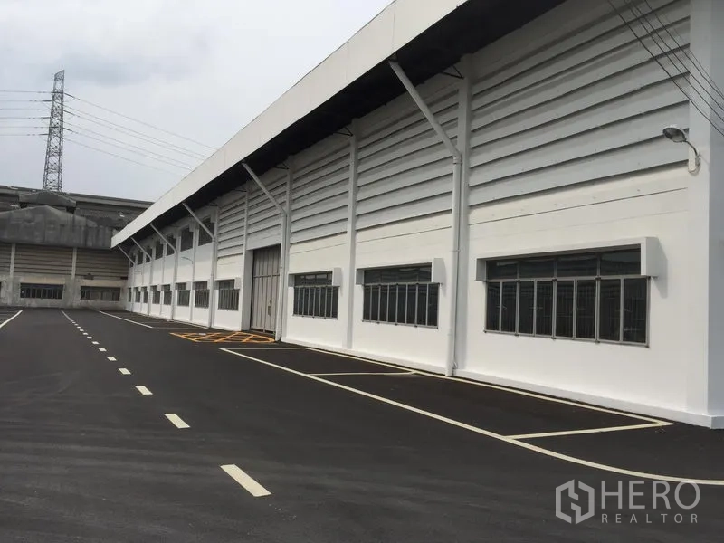Warehouse for rent in Lam Luk Ka, Pathum Thani - Modern white warehouse exterior with long loading bay, multiple windows, and a wide asphalt driveway with marked parking in Lam Luk Ka, Pathum Thani.