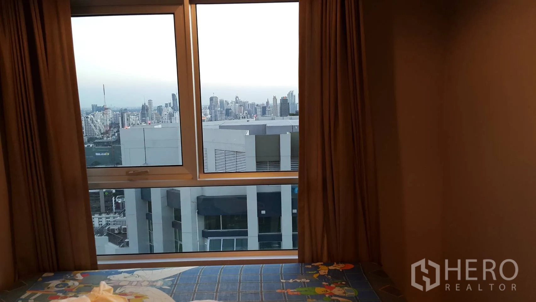 Condominium for rent in Huai Khwang, Bangkok - Second bedroom window framing Bangkok skyline and nearby building rooftops.