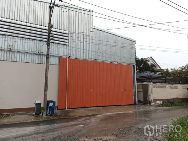 คลังสินค้า ให้เช่าในKhlong Sam Wa, Bangkok - ด้านหน้าของโกดัง ประตูบานเลื่อนสีส้มและผนังกัลวาไนซ์ ใกล้เขตรอยต่อบ้านพักอาศัย