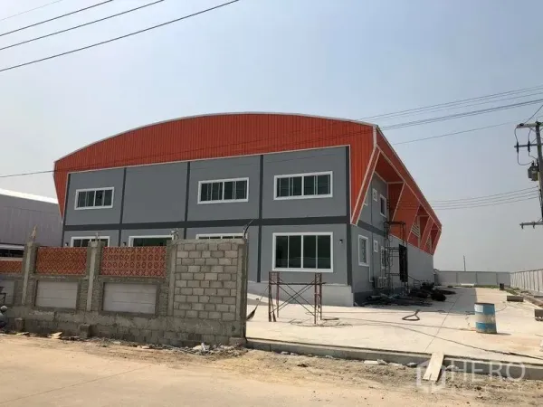 工厂 出售在Mueang Samut Sakhon, Samut Sakhon - 工厂侧视图，橙色金属包板与大窗，宽阔混凝土场地及电力线路。