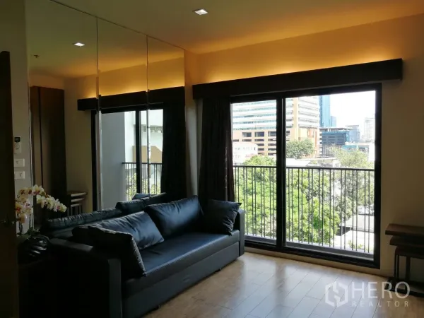 คอนโด ให้เช่าในWatthana, Bangkok - ห้องนั่งเล่นสว่าง โซฟาสีดำ กระจกเต็มบาน เปิดสู่ระเบียงวิวต้นไม้และเมือง