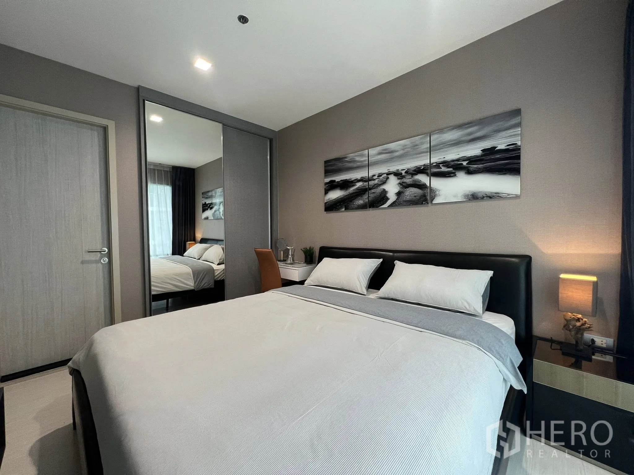 Condominium for rent in Khlong Toei, Bangkok - Cozy bedroom with upholstered bed, bedside lamp, desk, and sliding mirrored closet doors.