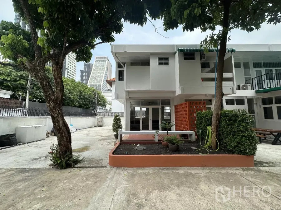 Home Office for rent in Watthana, Bangkok - Two-storey white home office with planters and mature trees, city skyline in the background.