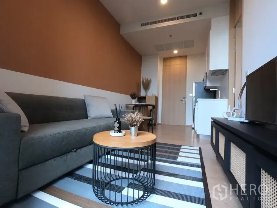 Condominium for rent in Watthana, Bangkok - Modern living room with grey sofa, round wooden coffee table, striped rug, TV console and open-plan layout toward the kitchenette.