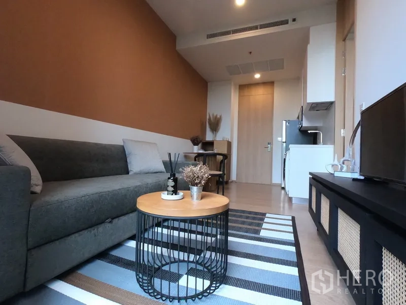 Condominium for rent in Watthana, Bangkok - Modern living room with grey sofa, round wooden coffee table, striped rug, TV console and open-plan layout toward the kitchenette.