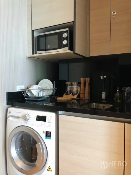 Condominium for rent in Khlong Toei, Bangkok - Built-in kitchenette featuring microwave, black countertop, sink, and front-loading washer.