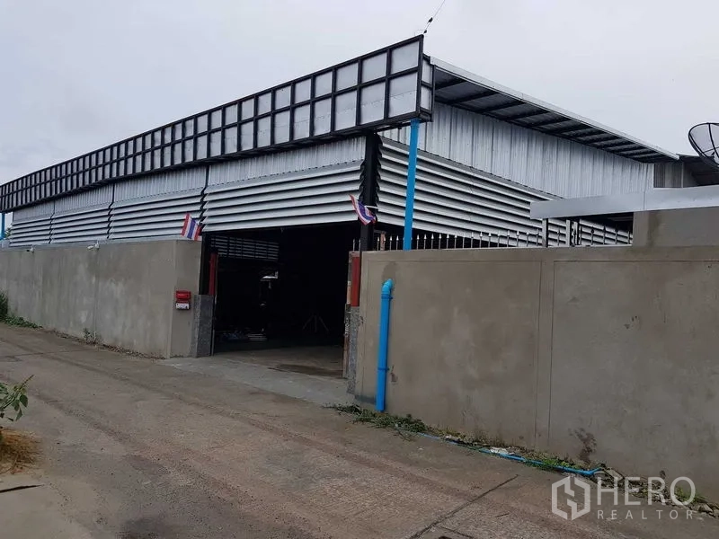 货栈 出售在Lam Luk Ka, Pathum Thani - 金属板外立面的仓库外景，主大门开启，蓝色排水管与高围墙可见。