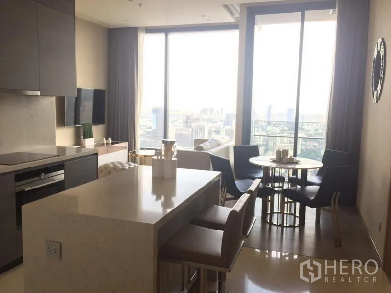 Condominium for rent in Watthana, Bangkok - Open‑plan kitchen with island, TV wall, dining set and large windows showcasing a city skyline view.