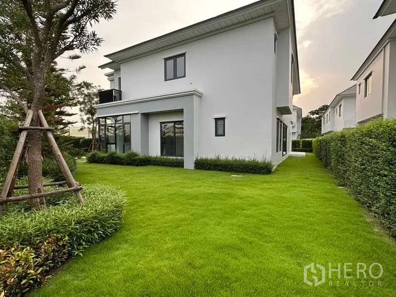 บ้าน ขายในLat Krabang, Bangkok - บ้านเดี่ยวสีขาวสองชั้นพร้อมสนามหญ้ากว้างและแนวรั้วพุ่มไม้ตัดแต่งเรียบร้อยยามเย็น