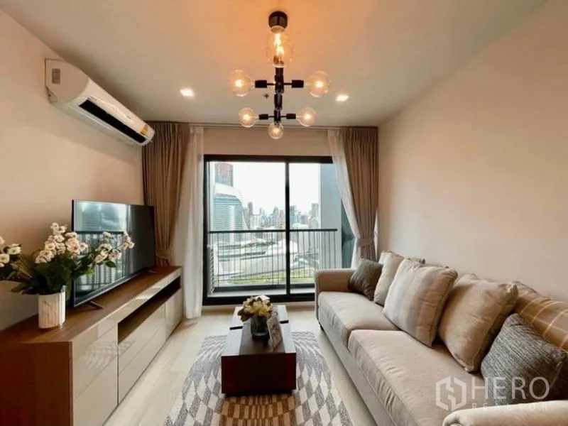 Condominium for rent in Pathum Wan, Bangkok - Living room with TV console, air-conditioner, chandelier, and sliding doors to a city-view balcony.