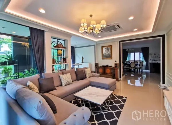 Detached House for rent or sale in Bang Kapi, Bangkok - Elegant living room with L-shaped sofa, chandelier lighting, marble coffee table, and view to dining area.