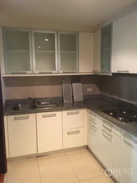 Condominium for rent or sale in Watthana, Bangkok - Compact U-shaped kitchen featuring white cabinetry, granite countertops, twin sinks and a built-in cooktop.