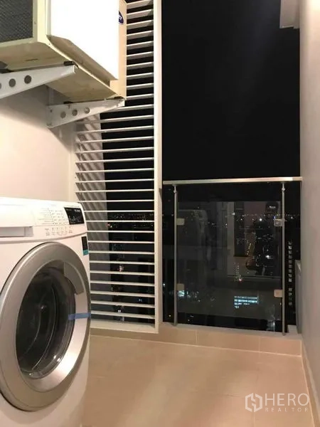 คอนโด ให้เช่าหรือขายในRatchathewi, Bangkok - ระเบียงครัวซักล้างพร้อมเครื่องซักผ้า ซี่บังลม และทิวทัศน์เมืองยามค่ำผ่านราวกระจก