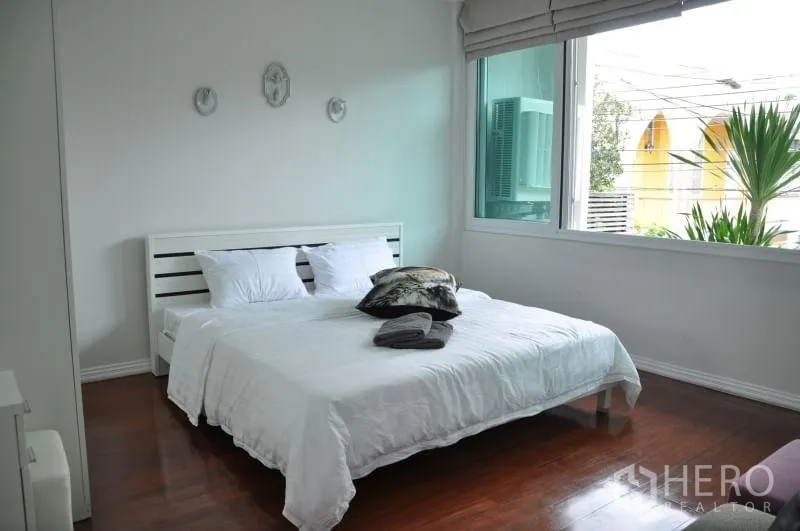 Townhouse for rent or sale in Watthana, Bangkok - Sunlit bedroom with double bed, white walls and wood flooring beside a large window.