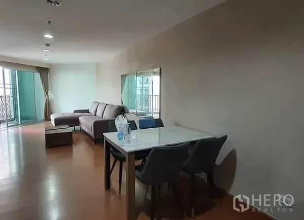 Condominium for rent in Huai Khwang, Bangkok - Open-plan living and dining space with a marble-top table and sliding door to a balcony with curtains.