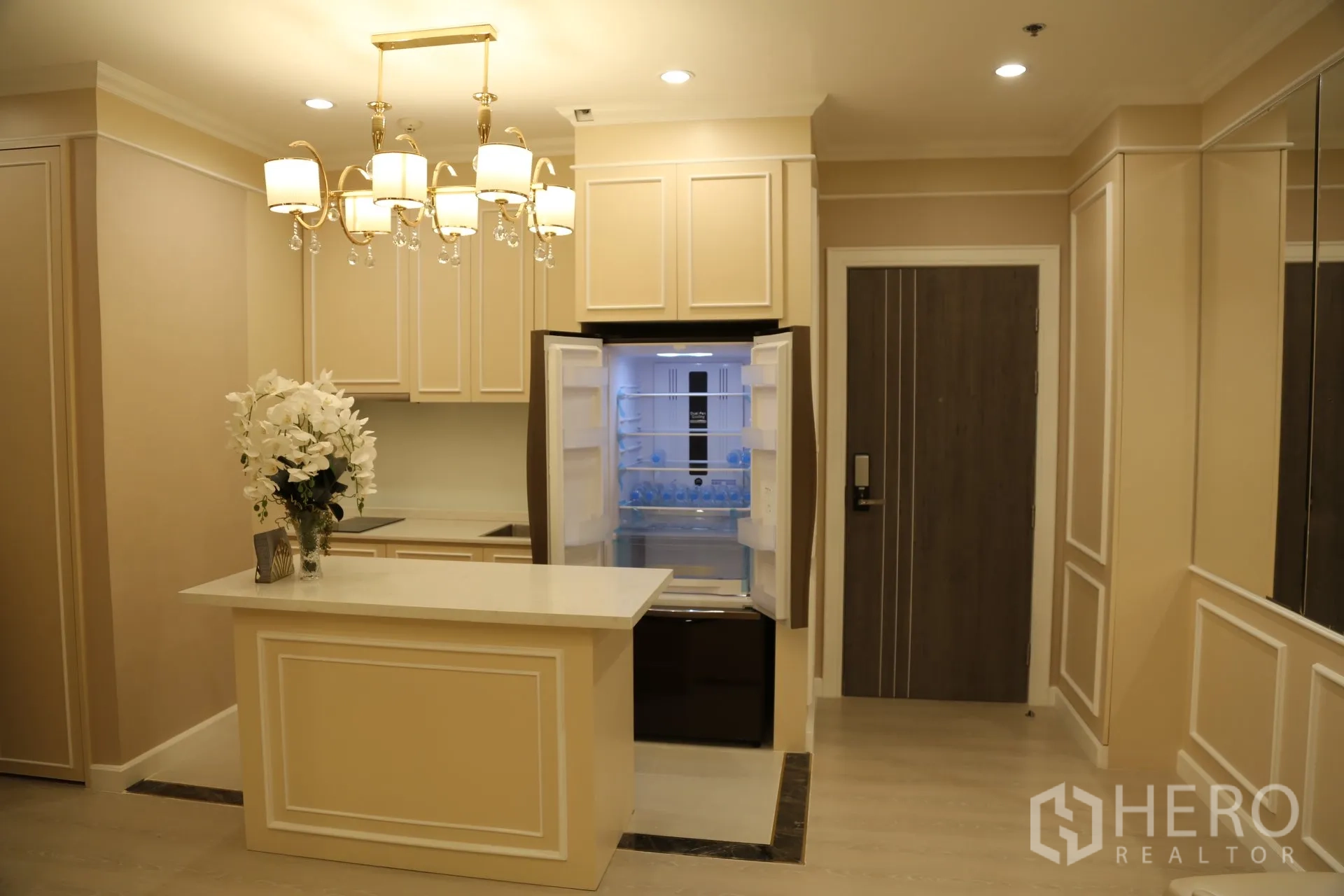 Condominium for rent in Huai Khwang, Bangkok - Modern beige kitchen with paneled cabinetry, island counter, pendant lighting and an open double-door refrigerator near the entry.